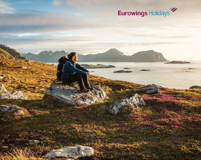 Eine Frau sitzt auf einem Felsen, der von einer skandinavischen Landschaft umgeben ist und schaut auf das weite Meer.