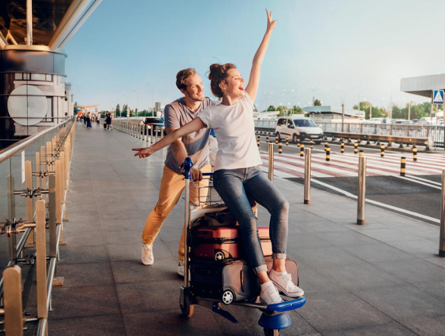 Ein Mann schiebt eine Frau vor dem Flughafen auf einem Kofferwagen,